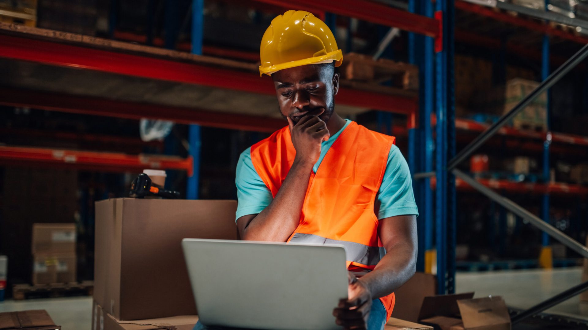Focused african american storekeeper sitting on boxes in warehouse and using laptop for checking inventory, he is thinking about solution for a problem