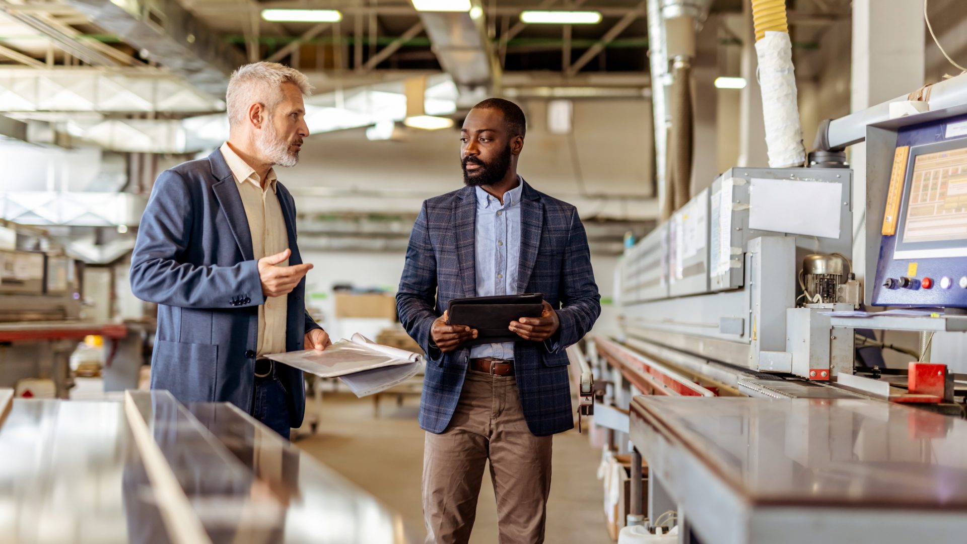 Two professionals interact in a manufacturing facility, discussing operations and reviewing data using modern equipment, fostering effective communication and teamwork in a professional industrial environment.