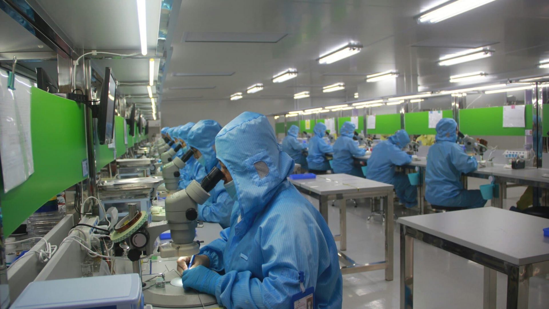 High-tech manufacturing cleanroom with workers in blue protective suits using microscopes for precision assembly.